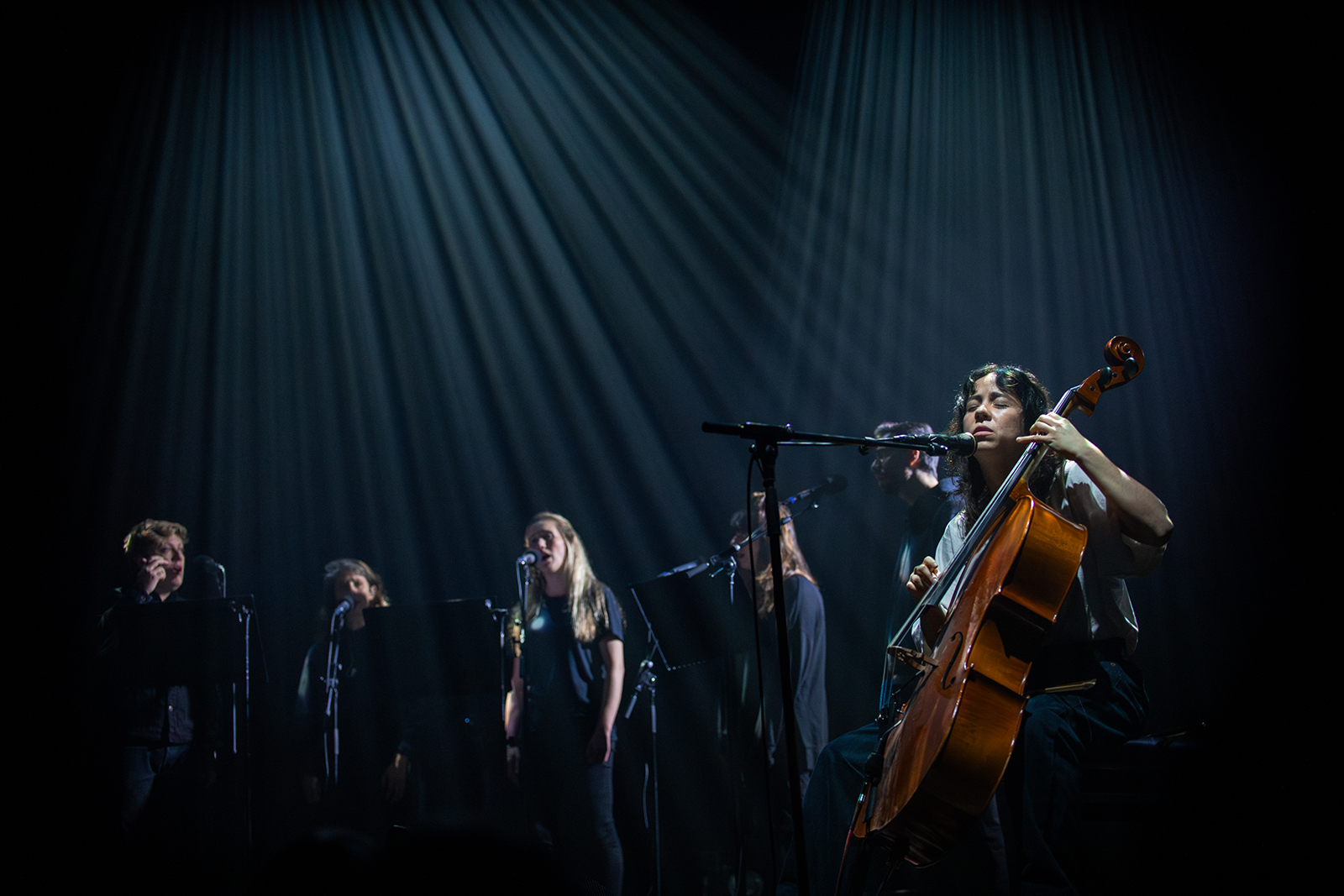 This is a photo of Mabe Fratti and Shards performing together. It is a dark photo in which streaks of light shine on the performers from above. On the left, the five artists of Shards are singing behind their microphones. On the right, Mabe Fratti sits with her cello.